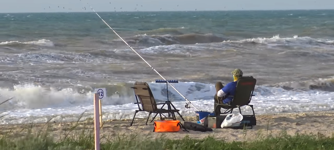 Рыбак сидит на кресле на берегу моря со снастями 