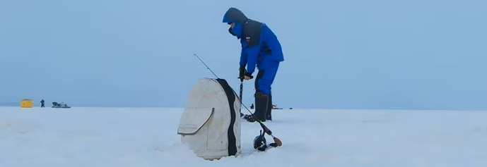 Рыбак в синем костюме делает лунку во льду ледобуром